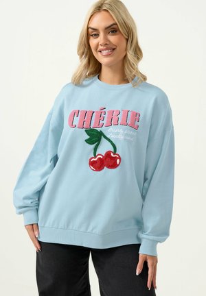 Frau mit blondem Haar, die einen hellblauen Pullover mit pinkem Schriftzug "CHÉRIE" und zwei roten Kirschen bestickt auf der Vorderseite trägt, lächelnd.