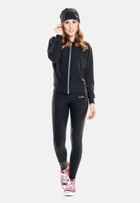 Black zip-up jacket, black leggings, pink high-top sneakers, and a black beanie. Smooth fabric with fitted design and minimalist logo detail.
