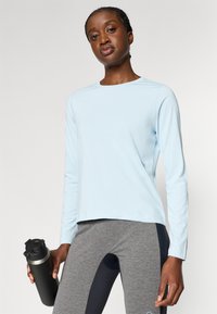 Hele sinine pika varrukaga sportlik särk, millel on mugav lõige, koos hallide retuusidega. Mudel hoiab käes musta veepudelit hõbedaste detailidega.