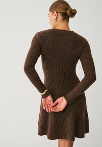 Frau mit einem Dutt, die goldene Creolen trägt und ein braunes geripptes Strickkleid anhat, mit den Händen hinter dem Rücken vor einem schlichten Hintergrund steht.