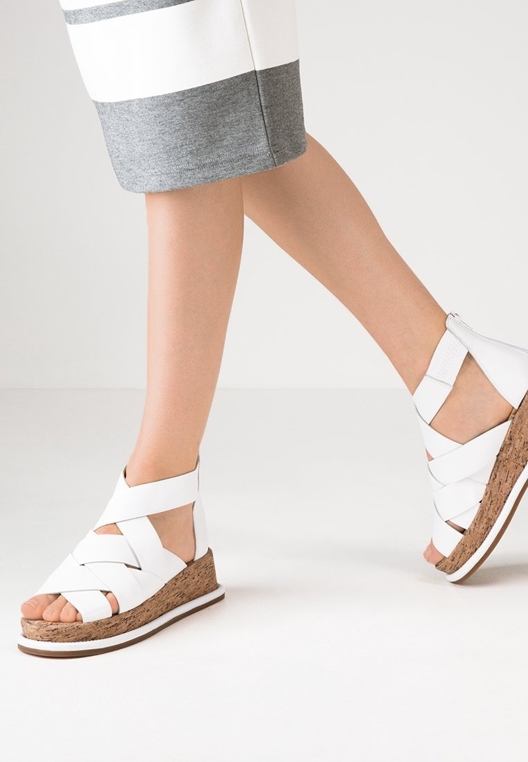 Jambes portant des sandales compensées en cuir blanc avec semelles en liège et brides autour de la cheville, associées à une jupe blanche et grise longueur genou.
