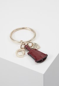 Porte-clés avec un anneau métallique doré et deux breloques : un mini sac en cuir bordeaux et une lettre "G" dorée, accompagnés d'une étiquette en forme de cœur dorée.