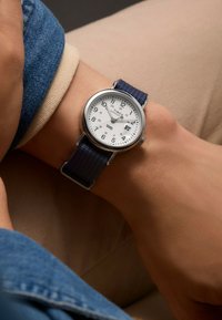 Timex watch with a silver bezel, white dial, black Arabic numerals, and a navy fabric strap, displayed on a wrist against a neutral background.