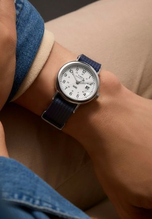 Montre Timex avec une lunette argentée, un cadran blanc, des chiffres arabes noirs et un bracelet en tissu bleu marine, présentée sur un poignet contre un fond neutre.