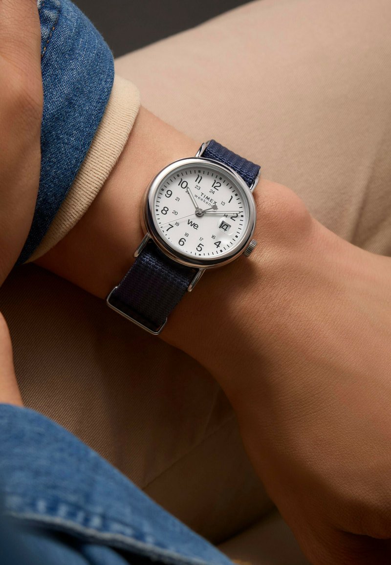 Timex sat sa srebrnim okvirom, bijelim brojčanikom, crnim arapskim brojevima i tamnoplavim tekstilnim remenom, prikazan na zapešću protiv neutralne pozadine.
