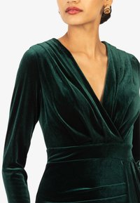 Groene fluwelen jurk met een diepe V-hals en lange mouwen. Heeft een getailleerde taille en een gedrapeerde corsage met een zachte textuur, wat zorgt voor een luxe uitstraling.