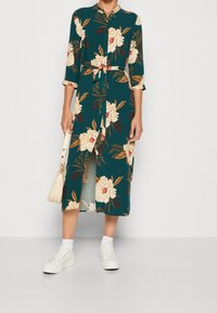 Abito midi floreale verde con colletto, maniche lunghe, chiusura frontale con bottoni e vita legata. Abbinato a sneaker bianche e una borsa color crema.