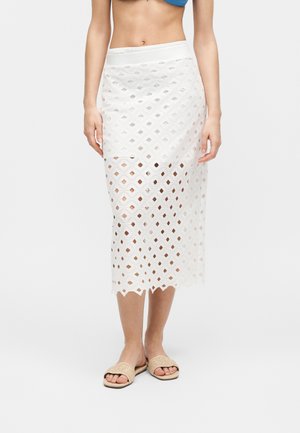 Žena nosí bielu midi sukňu s geometrickým vzorom výrezov cez jednofarebný spodný diel, doplnenú béžovými otvorenými sandálmi.