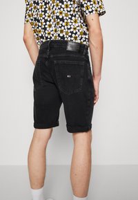 Pantalones cortos de mezclilla negros con dobladillos enrollados, que cuentan con un parche con logo en la parte trasera, diseño de cinco bolsillos y una estética minimalista y aerodinámica.