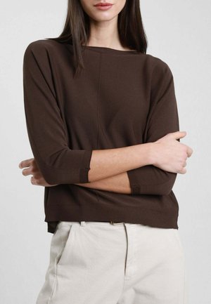Donna con capelli lunghi e scuri, vestita con una maglia marrone a maniche lunghe e pantaloni beige chiaro, in piedi con le braccia incrociate davanti a uno sfondo uniforme.