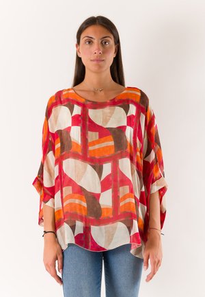 Donna con capelli lunghi e scuri che indossa una maglia larga con motivo geometrico rosso, arancione, marrone e bianco, abbinata a jeans blu, in piedi su sfondo bianco.