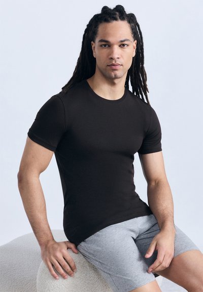 Νέος άνδρας με μακριά dreadlocks, φορώντας εφαρμοστό μαύρο μπλουζάκι και γκρι σορτς, κάθεται σε ένα υφασμένο στρογγυλό σκαμπό μπροστά σε απλό φόντο.