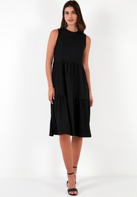 Black sleeveless dress with a tiered design, made of soft fabric. Features a high neckline and falls below the knee. Simple and versatile.