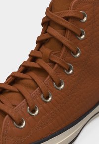 Καφέ αθλητικό παπούτσι από δερματίνη με αντίστοιχα κορδόνια περασμένα από ασημένιες τρύπες και λευκή ελαστική σόλα με μαύρο περίγραμμα.