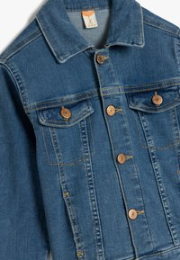 Chaqueta de denim de color azul, con dos bolsillos en el pecho con botones en tono dorado, un collar clásico y detalles de costura visibles.
