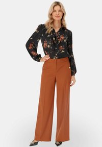 Kobieta w czarnej kwiatowej bluzce z kokardą i szerokich, rdzawych spodniach, stojąca z jedną ręką na biodrze na białym tle.