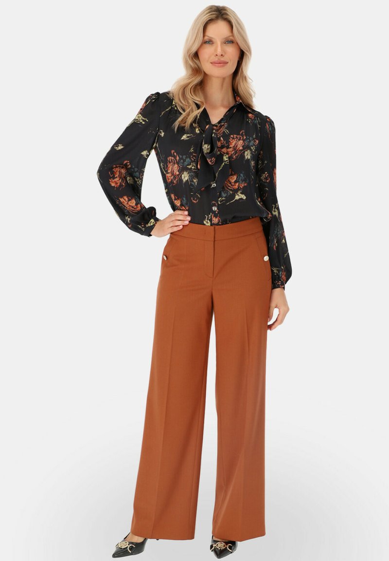 Kobieta w czarnej kwiatowej bluzce z kokardą i szerokich, rdzawych spodniach, stojąca z jedną ręką na biodrze na białym tle.