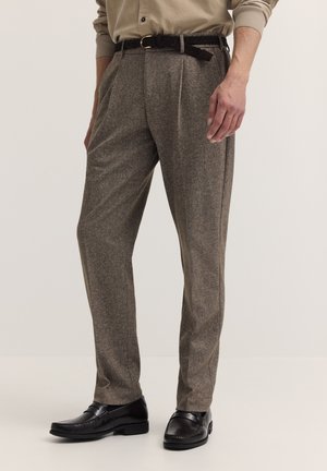 Mann trägt braune Wollhose mit schwarzem geflochtenem Gürtel und schwarzen Lederschuhen vor neutralem Hintergrund.