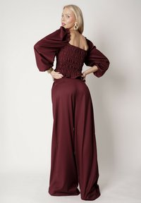 Burgundowy kombinezon z bufiastymi rękawami i marszczoną górą, posiadający szerokie, luźne nogawki oraz kwadratowy dekolt z tyłu. Gładki, teksturowany materiał.