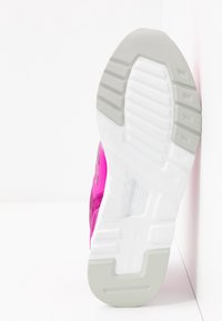 Semelle d'une chaussure de sport blanche et grise avec partie supérieure rose positionnée verticalement contre un coin formé par un mur blanc et le sol.