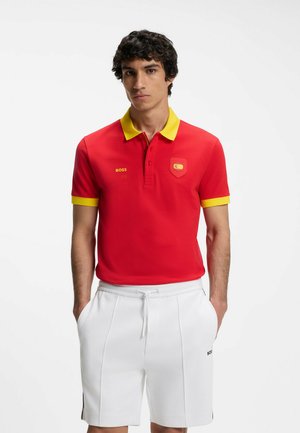 Man wearing red polo with yellow collar and sleeve trim, white drawstring shorts, standing with hands in pockets against white background.