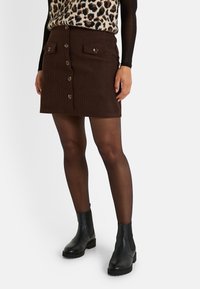 Falda de pana marrón con cierre de botones en el frente, dos bolsillos en el pecho con acentos de botones, combinada con botas negras hasta el tobillo y medias transparentes.