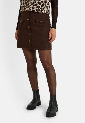 Falda de pana marrón con cierre de botones en el frente, dos bolsillos en el pecho con acentos de botones, combinada con botas negras hasta el tobillo y medias transparentes.