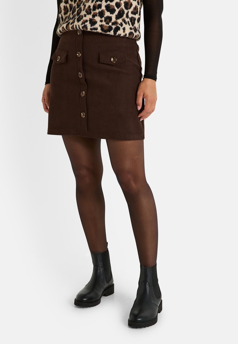Jupe en velours côtelé marron avec fermeture à boutons sur le devant, deux poches poitrine avec des accents de boutons, associée à des bottines noires et des collants transparents.