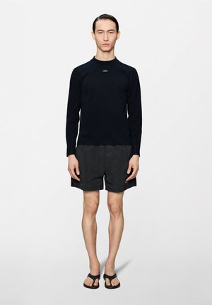 Férfi áll, fekete hosszú ujjú pólót, fekete rövidnadrágot és fekete szandált visel, egyszínű fehér háttér előtt.