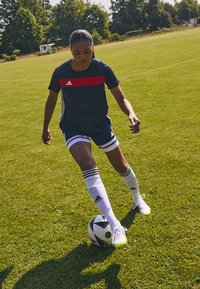 Adidas sportset bestaande uit een marineblauwe shirt met een rode streep, witte shorts en gestreepte witte sokken, op een grasvoetbalveld, een bal schoppend.