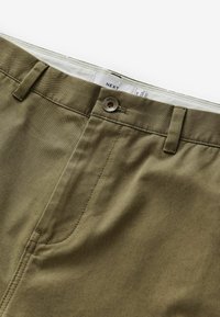 Pantalons en coton kaki avec une texture lisse, dotés d'une fermeture à bouton à l'avant, de passants de ceinture et de surpiqûres ton sur ton. Étiquette visible à l'intérieur de la taille.