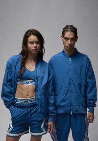 La ropa deportiva azul incluye una chaqueta bomber, un sujetador deportivo y pantalones cortos. La tela es suave y tiene un ligero brillo, con detalles de logotipos y acabados en contraste.