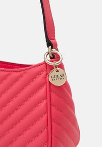 Rosa quiltet håndtaske med et guld rundt skilt, der har "GUESS EST 1981." Inkluderer en kontrastfarvet sort rem og en glat tekstur.