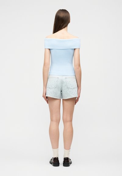 Γυναίκα με μακριά καστανά μαλλιά, γυρισμένη με την πλάτη, φοράει ανοιχτό μπλε μπλούζα με ακάλυπτους ώμους, ανοιχτόχρωμο τζιν σορτς, λευκές κάλτσες και μαύρα παπούτσια.