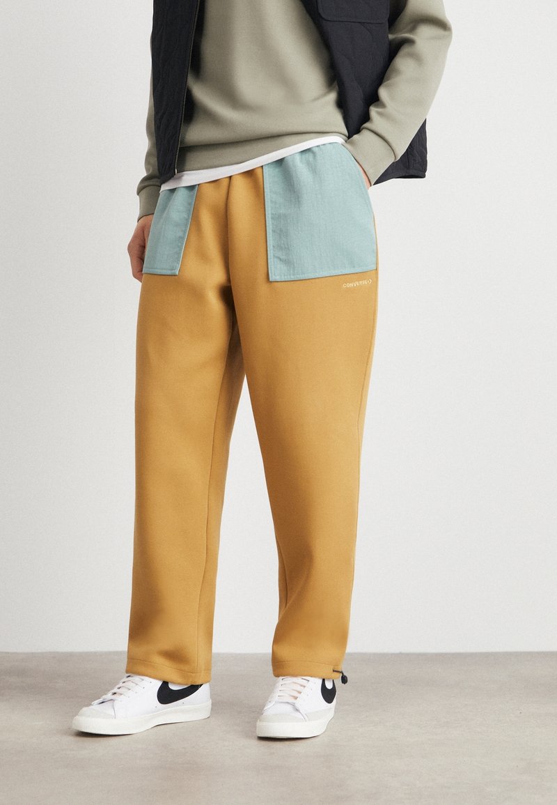 Jogginghosen in Senfgelb mit kontrastierenden hellblauen Patches und einem gestickten Logo. Der Stoff ist glatt und leicht texturiert. Die Schuhe sind weiß mit schwarzen Akzenten.