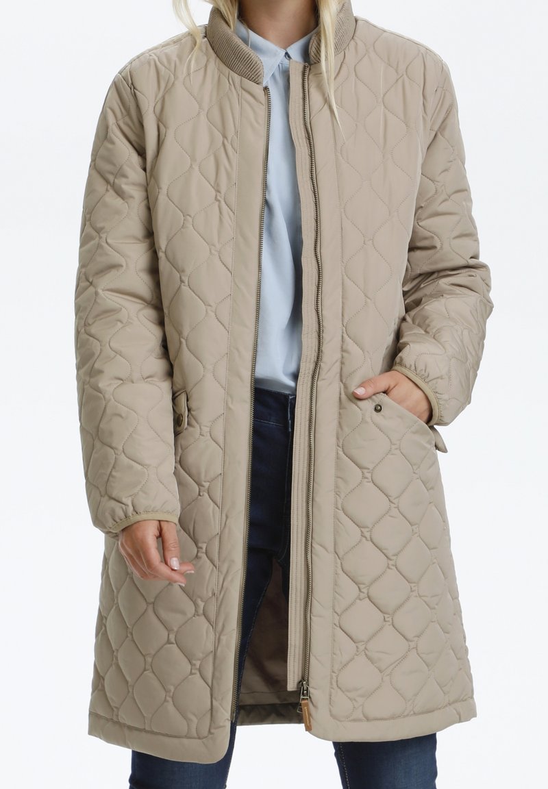 Beige quiltet jakke med høj krave, lange ærmer og lynlås foran. Har to sidelommer og en glat overflade.