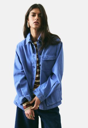 Junge Frau mit langen dunklen Haaren, die große Creolen trägt, eine blaue Jacke über einem gestreiften Hemd, dunkle Hosen und goldene Ringe an den Fingern.