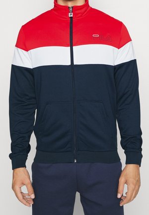 Mężczyzna noszący kurtkę zapinaną na zamek z poziomymi blokami kolorów w czerwieni, bieli i granacie oraz logo FILA na piersi.