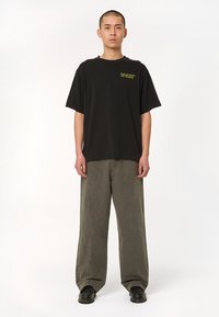 Black cotton t-shirt with short sleeves and a crew neck, featuring a small green graphic on the chest. Paired with loose, olive green pants.