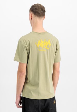 Man facing away wearing light green T-shirt with yellow "Alpha Industries" logo on the upper back and black pants with a patch.