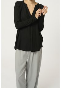 Kvinde iført en sort langærmet V-hals trøje og lysegrå bukser, som justerer ærmet med hænderne.