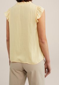 Femme portant un haut sans manches de couleur jaune pâle avec des épaules froncées et un pantalon beige, vue de dos sur un fond neutre.