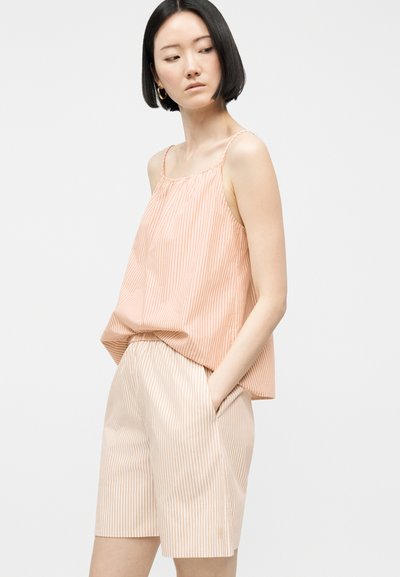 Γυναίκα που φοράει ριγέ αμάνικη μπλούζα με κάθετες ρίγες σε ροδακινί και άσπρο χρώμα και αντίστοιχα σορτς μέχρι το γόνατο, στέκεται με τα χέρια στις τσέπες.