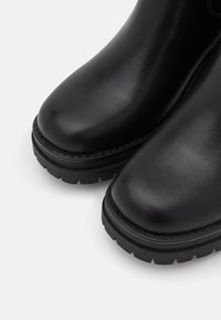 Bottes en cuir noir à finition lisse, bout arrondi et semelle en caoutchouc robuste avec des crampons profonds pour l'adhérence. Aucun élément métallique visible.