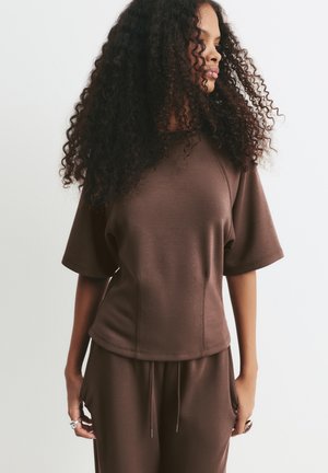 Woman with curly hair wearing a brown short-sleeve top and matching pants, standing against a plain white background.