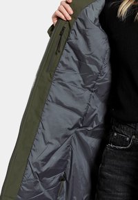 Interior de chaqueta verde oliva con forro gris, que cuenta con un bolsillo con cremallera y una textura suave y acolchada. Abierta para una clara visibilidad.