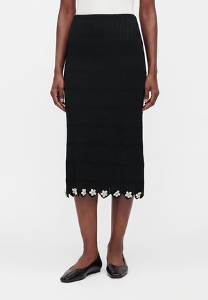 Jupe midi texturée noire avec ourlet festonné et ornements floraux blancs, portée avec des ballerines noires et un haut blanc sans manches.