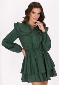 Grünes Baumwollkleid mit Rüschensaum, langen Ärmeln und gestuftem Rock. Dekorative Besätze entlang der Kanten. Knopfleiste vorne.