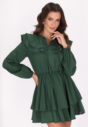 Groene katoenen jurk met een geplooid kraagje, lange mouwen en een gelaagde rok. Decoratieve accenttrim langs de randen. Knopsluiting aan de voorkant.
