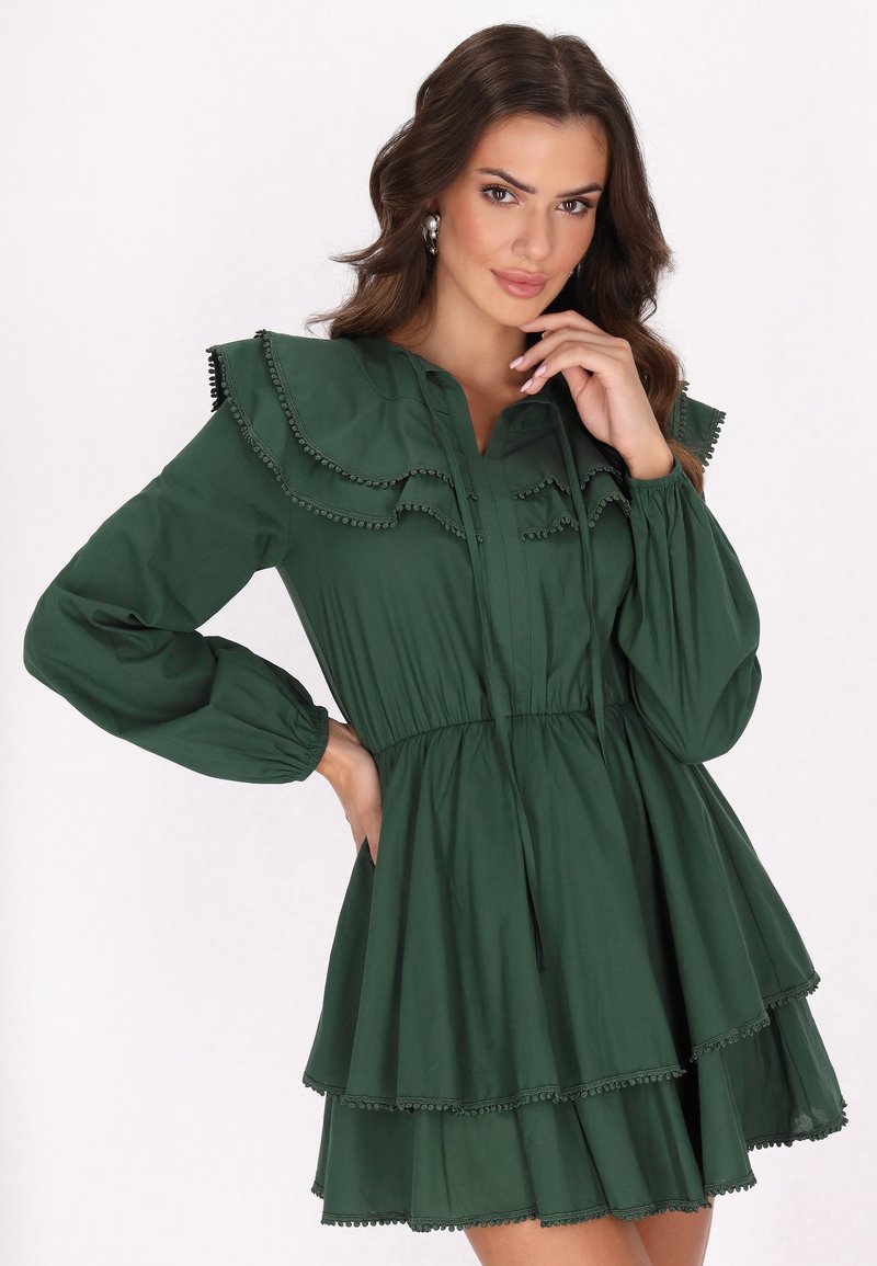Grünes Baumwollkleid mit Rüschensaum, langen Ärmeln und gestuftem Rock. Dekorative Besätze entlang der Kanten. Knopfleiste vorne.